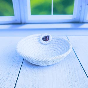 May include: A small, white, woven rope bowl with a dark brown button detail. The bowl is round with a shallow depth and sits on a white wooden surface. The background features a window with a green, blurred view.