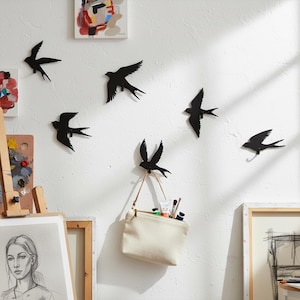 Op de afbeelding: Een verzameling zwarte metalen wanddecoraties in de vorm van zwaluwen in verschillende houdingen, bevestigd aan een witte muur. Eén zwaluw dient als haak en houdt een kleine canvas tas met kunstbenodigdheden vast. Kunstwerken en een ezel zijn zichtbaar op de achtergrond.