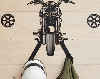 May include: Black metal motorcycle-themed wall art with the name "CLEMENT" above the handlebars. The art piece includes a motorcycle, two wheels, and two hooks holding a white helmet and a green jacket. The art is mounted on a light wood-paneled wall.