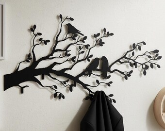 Puede incluir: Arte de pared de metal negro con un diseño de rama de árbol con hojas y tres pájaros. La rama tiene varios ganchos para colgar objetos. Una chaqueta negra cuelga de uno de los ganchos. El arte está montado en una pared blanca.
