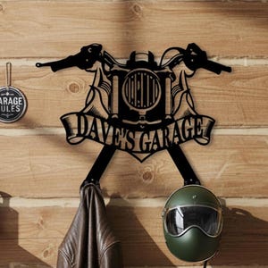 May include: Black metal motorcycle-themed wall decor with the text "DAVE'S GARAGE". The decor includes a motorcycle handlebar design, a helmet, and a leather jacket hanging below. A round sign with the text "GARAGE RULES" is also visible.