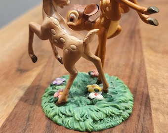 Puede incluir: Una figura de Disney que muestra dos cervatillos marrones con manchas blancas jugando sobre una base circular de hierba verde decorada con pequeñas flores de colores. Los cervatillos están en poses dinámicas, uno de pie y otro saltando, con la palabra DISNEY grabada en la base.
