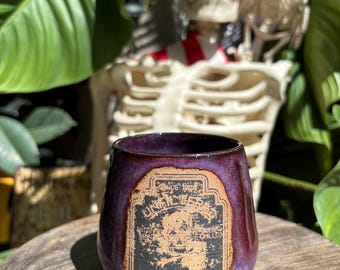 May include: A ceramic tumbler with a deep purple glaze and a vintage-style label that reads "UNFILTERED POISON". The label is in black and tan, with a skull graphic. The tumbler is on a wooden surface.