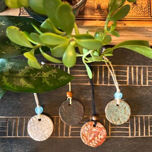 May include: Close-up of four handmade ceramic pendants with jute twine and wooden beads. The pendants are circular, with varying textures and colors, including white, brown, and orange with a floral design. Green plant leaves are in the background.