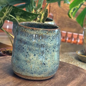 Puede incluir: Un vaso de cerámica hecho a mano con un esmalte moteado azul-verdoso. La pequeña taza cilíndrica tiene una base redondeada y un borde ligeramente ensanchado. La cerámica se exhibe sobre una bandeja de madera, con plantas verdes frondosas en el fondo.