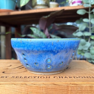 May include: A round ceramic berry bowl featuring a vibrant blue glaze with subtle green undertones. The bowl has a series of small, circular drainage holes spaced evenly around the lower half of its exterior, with a light-coloured, unglazed base.