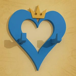 Puede incluir: Un colgador de pared azul en forma de corazón con una corona dorada. El corazón tiene dos ganchos y está montado en una pared amarillo claro. El diseño es una representación estilizada de un corazón y una corona.