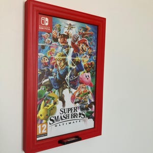 Può includere: Una custodia del gioco Super Smash Bros. Ultimate per Nintendo Switch incorniciata in una cornice rossa. La copertina del gioco presenta vari personaggi. Una cartuccia di gioco è visibile sotto. Il gioco è classificato 12.