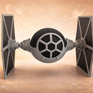 Puede incluir: Modelo detallado de una nave TIE fighter, con una cabina esférica central y dos grandes paneles solares angulados. El modelo es principalmente gris y negro, con detalles intrincados. El fondo es un degradado suave y borroso.
