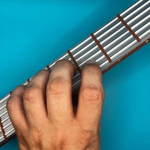 May include: A fretboard with silver frets and brown fret markers is held by a hand against a teal background. The frets are parallel and evenly spaced. The hand is positioned to play chords on the fretboard.