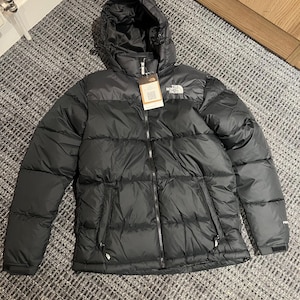 Könnte beinhalten: Schwarze Kapuzen-Steppjacke mit Reißverschluss und zwei Seitentaschen. Die Jacke hat das Markenlogo auf der Brust und ein Etikett am Reißverschluss. Die Jacke ist für kaltes Wetter konzipiert.