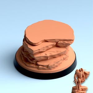 May include: A 3D-printed Hero Terrain model with the text "Hero Terrain STEPS IN THE DESERT". The model features a layered rock formation in a light orange color, set on a black base. A miniature figure stands on a smaller rock formation.