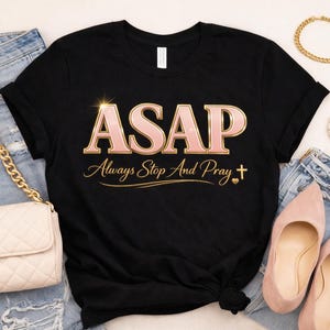 May include: Black t-shirt with the text "ASAP Always Stop And Pray" in pink and gold. The image includes a cream-coloured quilted handbag, blue jeans, pink shoes, and gold jewellery, including a bracelet and necklace.