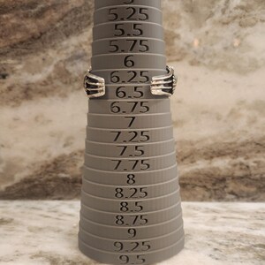 May include: A silver ring sizing tool with a grey plastic base. The tool has a series of rings that are marked with sizes from 5 to 9.5 in increments of 0.25.