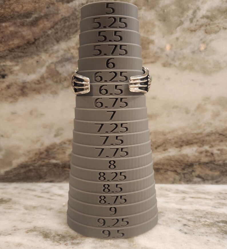 May include: A silver ring sizing tool with a grey plastic base. The tool has a series of rings that are marked with sizes from 5 to 9.5 in increments of 0.25.