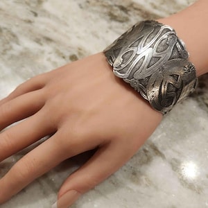 May include: A silver cuff bracelet with an intricate, embossed design. The bracelet features a detailed pattern of swirling lines and shapes. The metal has a slightly aged appearance. The bracelet is shown on a hand.