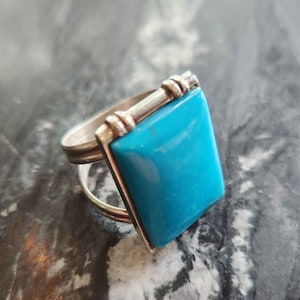 May include: A silver ring with a large, square, bright turquoise stone. The ring has a simple band with a decorative element connecting the stone to the band. The turquoise stone is the focal point.