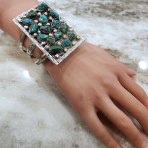 May include: A silver cuff bracelet featuring a rectangular face set with turquoise stones and small silver beads. The bracelet has three bands that wrap around the wrist. The bracelet is worn on a person's hand.