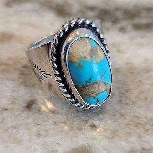 May include: A silver ring featuring a large, oval turquoise stone. The stone is a bright blue with brown markings. The ring has a twisted rope design around the stone and a sunburst pattern on the side.