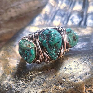 May include: A silver ring featuring three turquoise stones set in a twisted band. The stones are a deep green colour with brown veins.