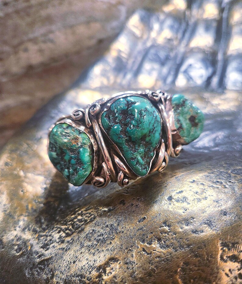 May include: A silver ring featuring three turquoise stones set in a twisted band. The stones are a deep green colour with brown veins.