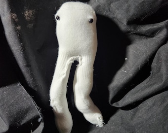 Può includere: Un burattino di calzino bianco con occhi a bottone neri e gambe lunghe. Il burattino è fatto da un calzino bianco e ha un aspetto semplice e fatto a mano.