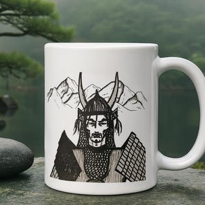May include: White ceramic mug featuring a black and white illustration of a samurai warrior with a horned helmet and mountain backdrop. The mug has a curved handle and is set against a blurred natural background.