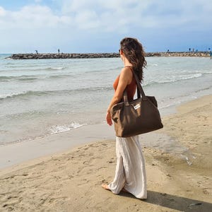 May include: A large, olive-green tote bag with two front pockets and a small white label. The bag has two long handles and is carried by a person on a sandy beach. The person is wearing a burnt orange top and white pants.