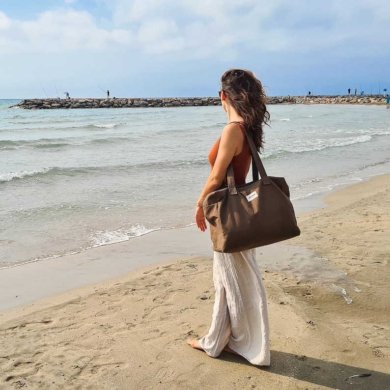 May include: A large, olive-green tote bag with two front pockets and a small white label. The bag has two long handles and is carried by a person on a sandy beach. The person is wearing a burnt orange top and white pants.