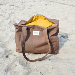 May include: A brown tote bag with a yellow interior, sitting on sand. The bag has long handles and a zipper closure. A white label with the text "L&Eacute;A PILEA" is visible on the front of the bag. The bag is open, revealing the yellow lining.
