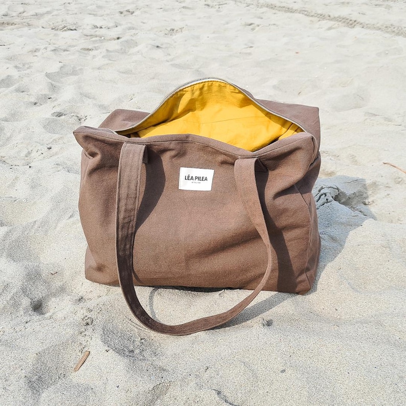 May include: A brown tote bag with a yellow interior, sitting on sand. The bag has long handles and a zipper closure. A white label with the text "L&Eacute;A PILEA" is visible on the front of the bag. The bag is open, revealing the yellow lining.