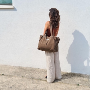 May include: A large, brown tote bag with two handles and a small white label is held by a person. The bag is made of a sturdy material and has a rectangular shape. The person is wearing a rust-colored top and cream-colored pants.