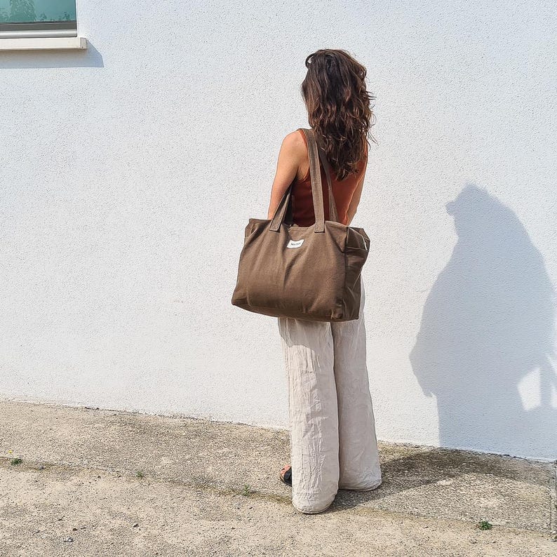 May include: A large, brown tote bag with two handles and a small white label is held by a person. The bag is made of a sturdy material and has a rectangular shape. The person is wearing a rust-colored top and cream-colored pants.
