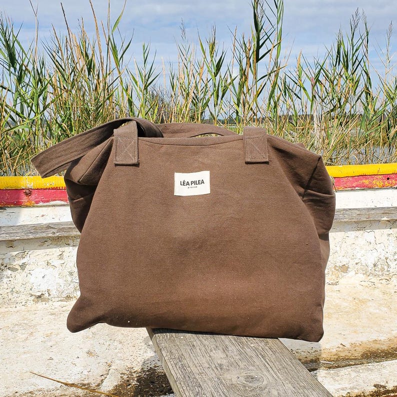 May include: A brown tote bag with a rectangular white label that reads "L&Eacute;A PILEA". The bag has two handles and a shoulder strap. The bag is made of a sturdy fabric and is sitting on a wooden surface.