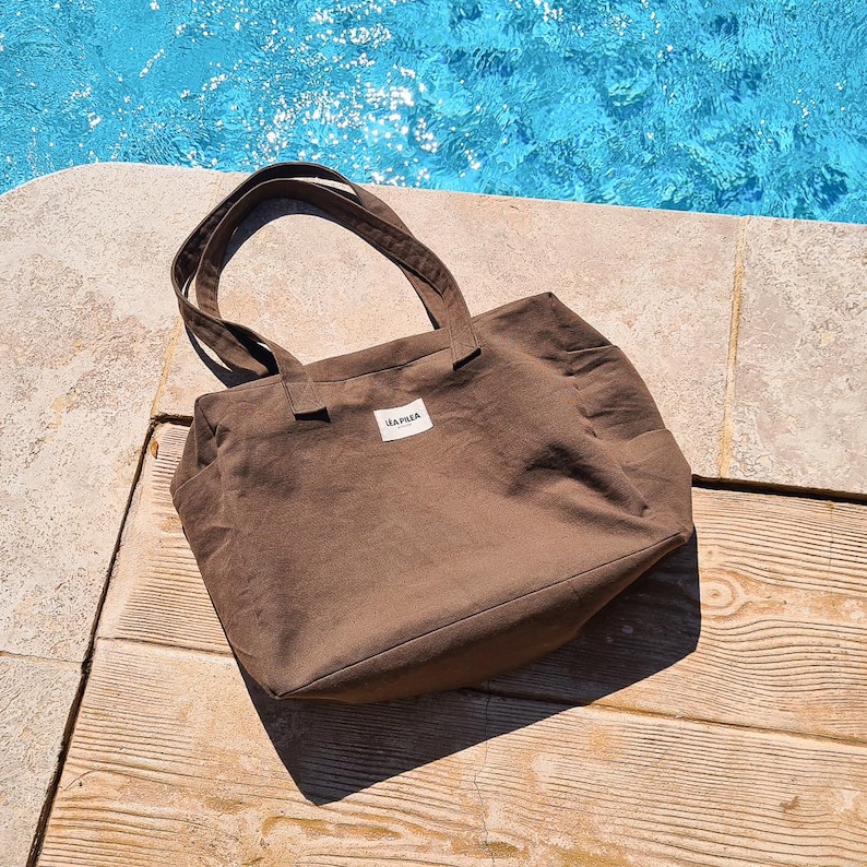 May include: A brown tote bag with two handles and a small rectangular label. The bag is made of a sturdy fabric and has a simple, functional design. The bag is sitting on a wooden surface.