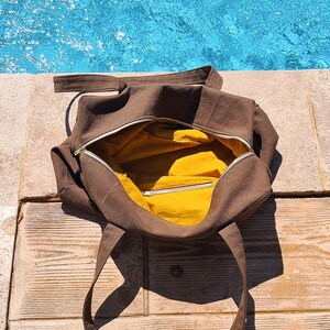May include: A brown tote bag with a yellow interior, open to reveal the inside. The bag has a silver zipper and long straps. The bag is sitting on a wooden surface near a pool.