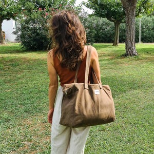 May include: A person is seen from behind, carrying a large olive-green tote bag with long handles. The bag has a rectangular shape and a small white label. The person is wearing a rust-colored tank top and cream-colored pants, standing in a grassy area.