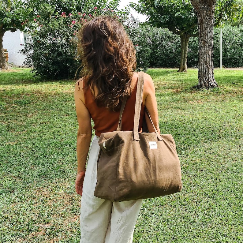 May include: A person is seen from behind, carrying a large olive-green tote bag with long handles. The bag has a rectangular shape and a small white label. The person is wearing a rust-colored tank top and cream-colored pants, standing in a grassy area.