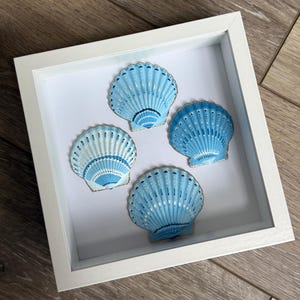 Op de afbeelding: Een vierkante witte schaduwdooslijst met vier individuele sint-jakobsschelpen op een witte achtergrond. Elke schelp is beschilderd in verschillende tinten licht- en donkerblauw met gedetailleerde witte stippenpatronen en fijne radiale ribbels.