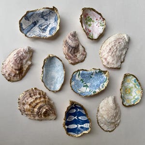 May include: A collection of decorative oyster shell dishes with gold gilded edges. The shells are lined with patterned paper in various designs, including nautical maps, floral prints, and fish motifs. The shells are arranged on a neutral surface.