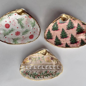 May include: Three decorative seashell dishes with gold rims. One dish features a white background with candy canes, fir branches, and red accents. Another has a pink background with green Christmas trees. The third has a cream background with a floral design.