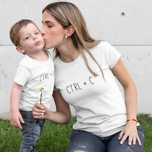 以下が含まれることがあります： 女性と子供が「CTRL +」と「CTRL + C」の文字が印刷された白いTシャツを着ています。女性は子供の頬にキスをしています。彼らは白い壁の前に立っており、手前に緑の草があります。