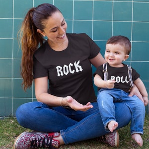 May include: A woman and a child wearing matching black t-shirts with white text. The woman's shirt says "ROCK" and the child's shirt says "ROLL". They are sitting on a green grassy surface in front of a blue tiled wall.