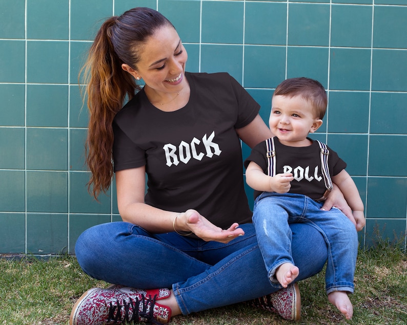 May include: A woman and a child wearing matching black t-shirts with white text. The woman's shirt says "ROCK" and the child's shirt says "ROLL". They are sitting on a green grassy surface in front of a blue tiled wall.