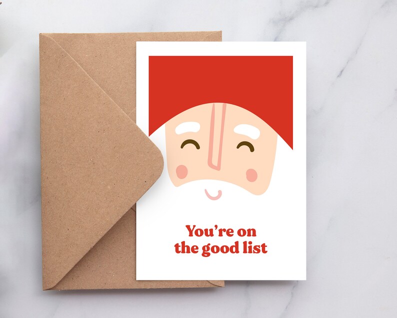 May include: A white greeting card featuring a stylized illustration of a face with a red hat, white eyebrows, and a white beard. The text "You're on the good list" is printed in red below the face. A brown kraft paper envelope is positioned behind the card.