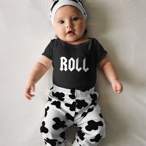 May include: A baby wearing a black bodysuit with the word "ROLL" printed on it, black and white cow print trousers, and a white hat with cow print ears.