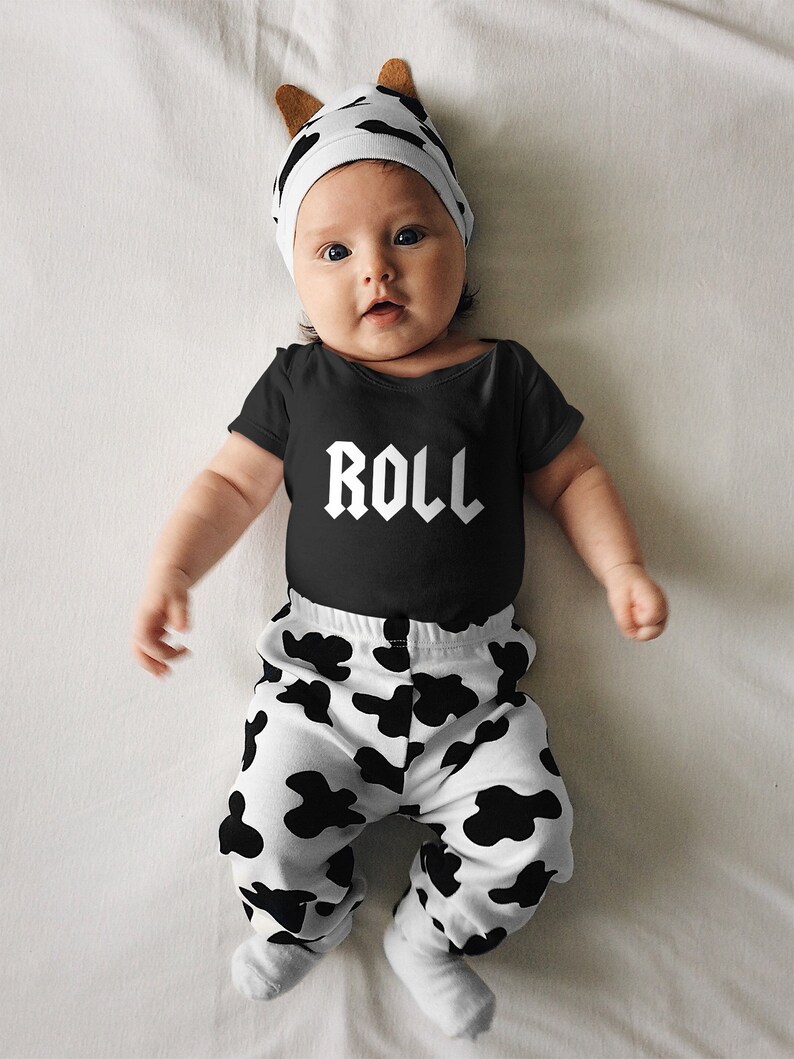 May include: A baby wearing a black bodysuit with the word "ROLL" printed on it, black and white cow print trousers, and a white hat with cow print ears.