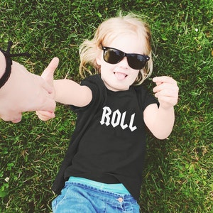 May include: A young child wearing a black t-shirt with the word "ROLL" printed in white on the front. The child is wearing sunglasses and is lying on their back in a grassy field.
