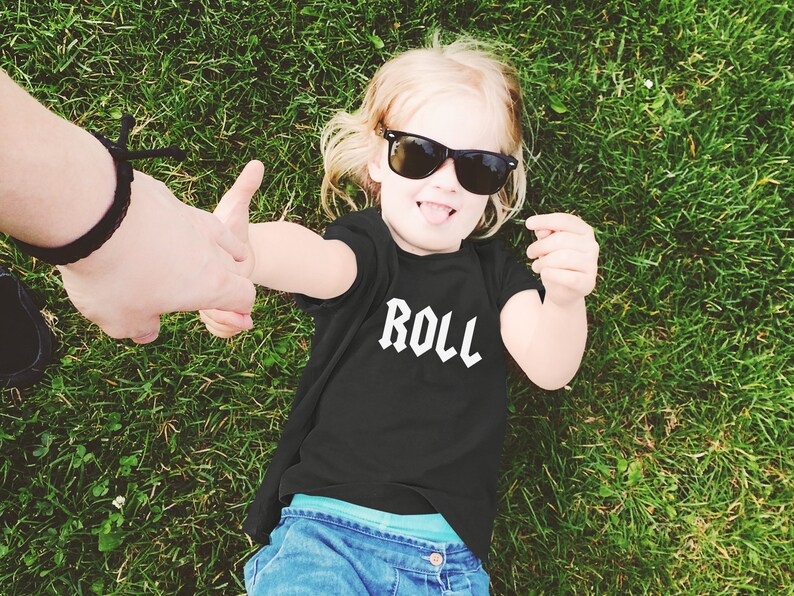 May include: A young child wearing a black t-shirt with the word "ROLL" printed in white on the front. The child is wearing sunglasses and is lying on their back in a grassy field.