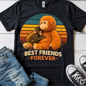 May include: Black t-shirt featuring a graphic of two monkeys hugging, set against a retro sunset design. The text "BEST FRIENDS = FOREVER =" is printed below the image. The shirt is paired with blue jeans and black sneakers.